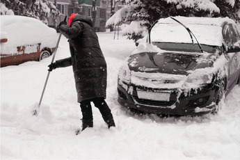 A person wearing a winter coat shovels snow near a snow-covered car.