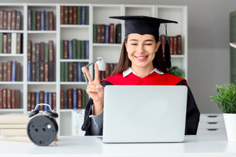 A young woman sitting at a desk in front of a laptop, wearing a college graduates cap and gown.