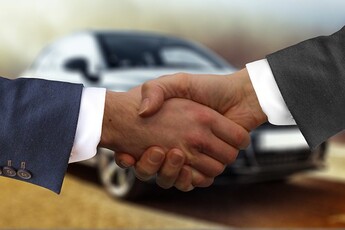 A man is shaking a car dealer's hand with a car in the background.