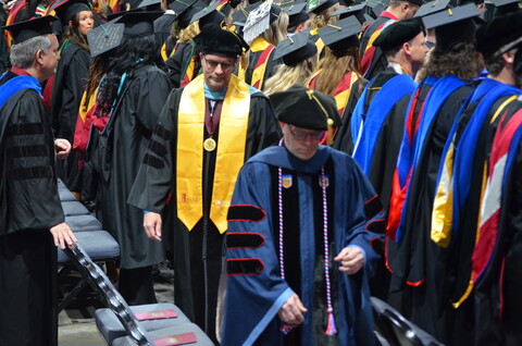 Many graduating students are standing in a room, proudly showing off their graduation gear. They have their caps and gowns on, some with gold and red tassels. They are all dressed in blue and red colors, which are common colors for graduation attire. The students are gathered in the room, ready to celebrate their achievements.