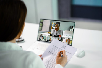A person is attending a video conference call on a laptop, with several participants visible on screen. The person is also holding a document and a pen, appearing to take notes.